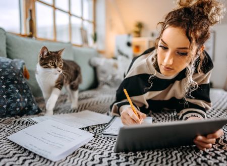 Eine junge Frau liegt entspannt mit ihrer Katze auf dem Sofa und liest in einem Tablet. Rundherum liegen handgeschriebene Notizen