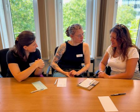 Drei Teilneherinnen unterhalten sich an einem Tisch, vor ihnen Notizzettel und Stifte.