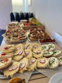 Auf einem Tisch stehen mehrere Tablets mit belegten Brötchen, Brezeln und Obst. Im Hintergrund mehrere Thermoskannen.